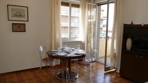 Een eetruimte met een ronde tafel gedekt voor vier personen, transparante stoelen, een houten vloer, een televisie op een kast en een balkon met een hekwerk.