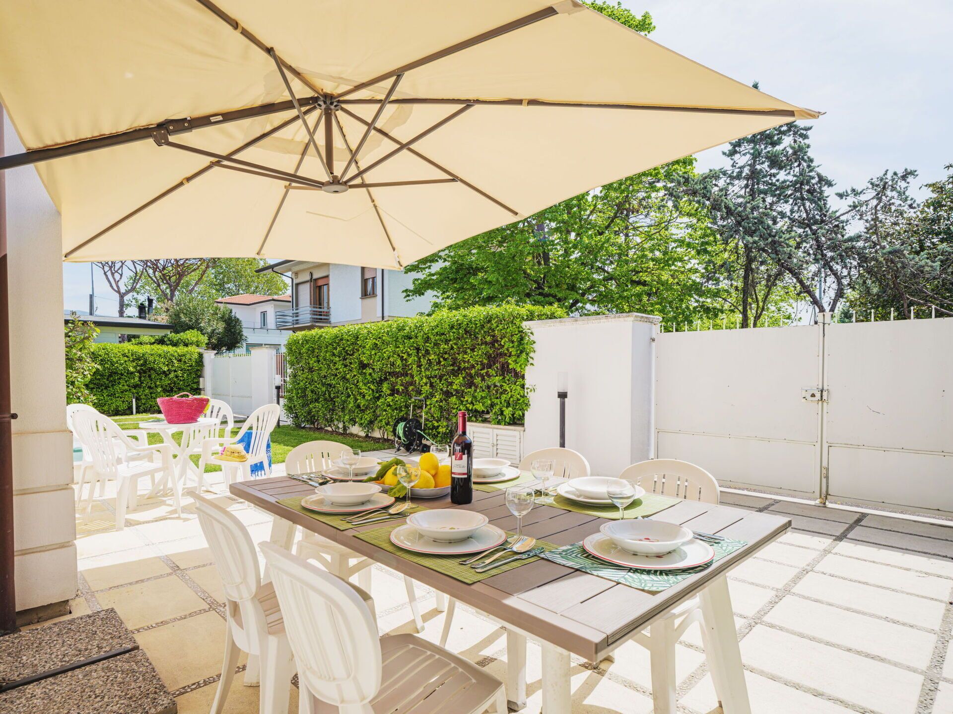Een terras buiten met een eettafel, omringd door witte stoelen, onder een grote parasol.