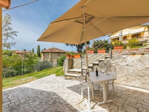 Uma área de pátio com mesa e cadeiras sob um grande guarda-sol, degraus de pedra e vista para uma colina com construções.
