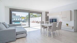 Een moderne woonkamer met een grijze bank, een eettafel met stoelen, een aan de muur bevestigde televisie en uitzicht op een zwembad en een terras met zitplaatsen.