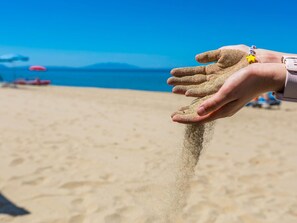 Uma mão segurando areia, com uma praia e guarda-sóis ao fundo.