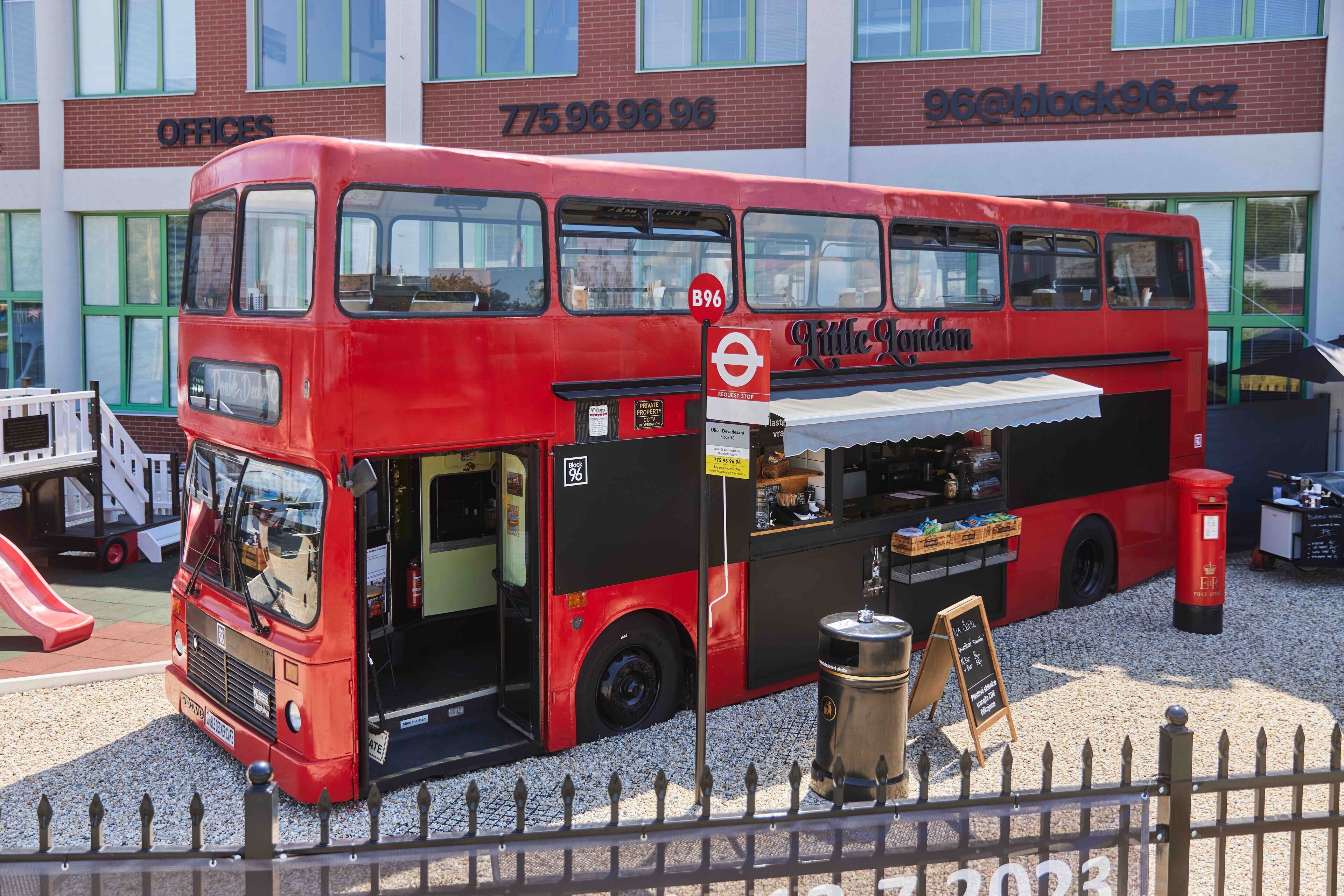 A red double-decker bus with a food stall attached, parked in front of a building with the sign "Offices" and contact number "775 96 96 96".