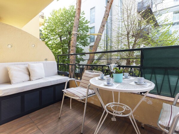 Un balcon avec un canapé blanc, une table ronde avec des tasses et un pot bleu, et une vue sur des immeubles et des arbres.