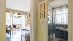 Uma sala de estar com área de jantar, uma porta de vidro que dá para uma varanda e uma cozinha com azulejos azuis e cortina de janela estampada.