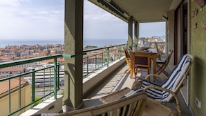 Varanda com mobiliário de vime, uma mesa e vista para a cidade e o mar.