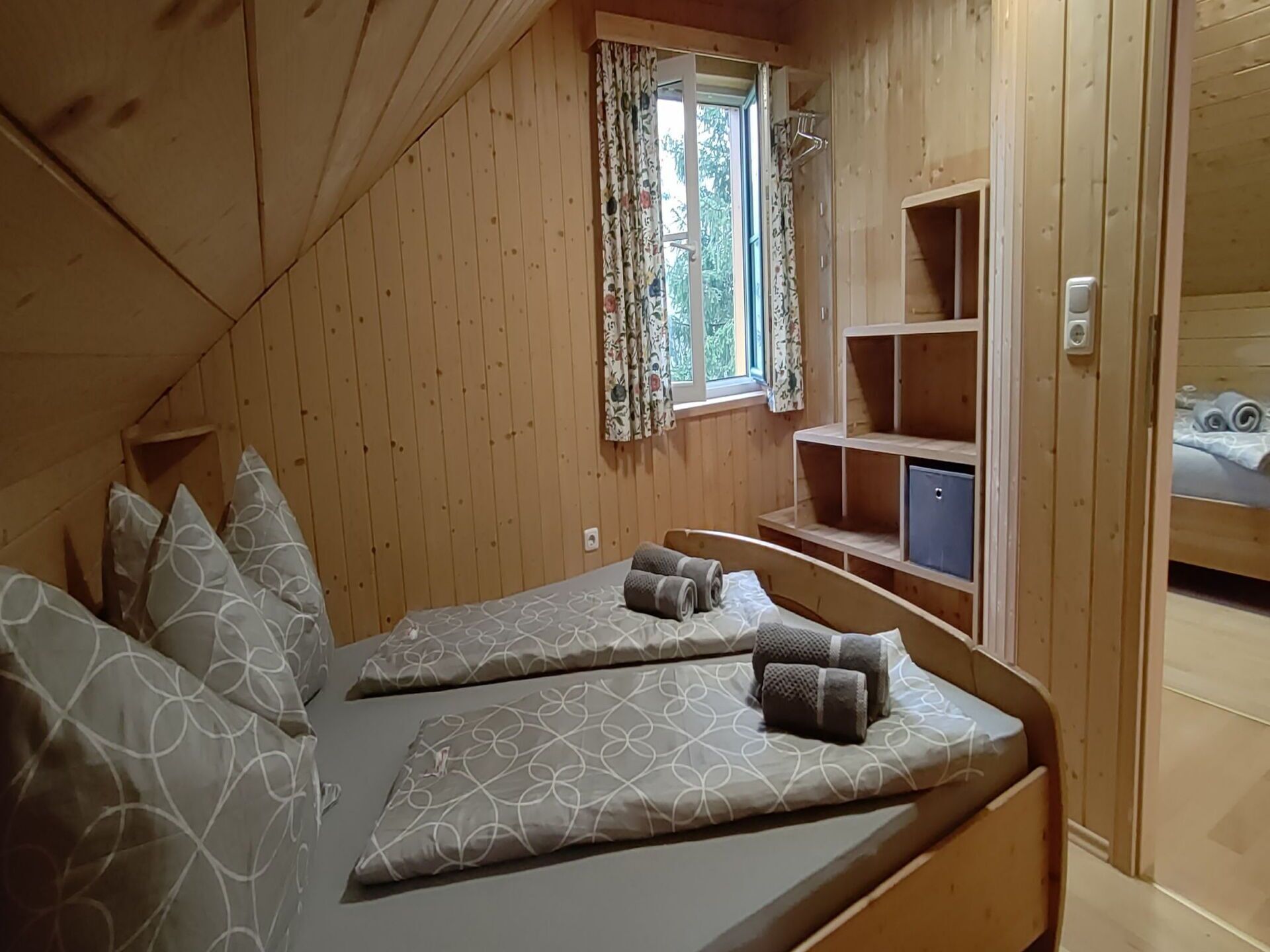 Een knus kamer in chaletstijl met twee bedden, een raam met bloemen gordijnen en inbouwplanken.