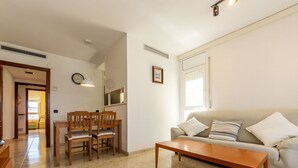 Un salón con sofá, una mesa de centro y una mesa de comedor con sillas. Hay una ventana con persianas, un reloj en la pared y una puerta que da a otra habitación.