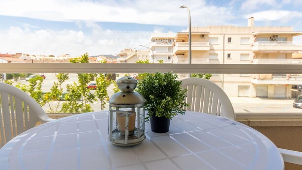 Mesa redonda con mantel blanco, un farol y una planta en maceta en un balcón con vistas a una zona residencial con edificios y una calle.