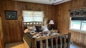 A wooden-paneled room with a bed featuring a quilt, pillows with motivational phrases, a nightstand with a lamp, and a window with a view. - Make memories at this Big Chetac Lake Cottage (Birchwood)