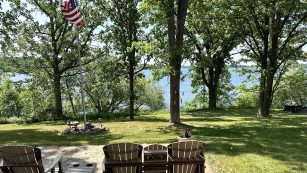 A lakeside outdoor seating area with Adirondack chairs and a table, a flag, and a view of the lake. - Make memories at this Big Chetac Lake Cottage (Birchwood)