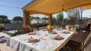 Une terrasse extérieure dotée d’un espace repas couvert, de piliers en pierre et d’une table dressée pour quatre personnes, avec des serviettes orange et un panier de fruits.
