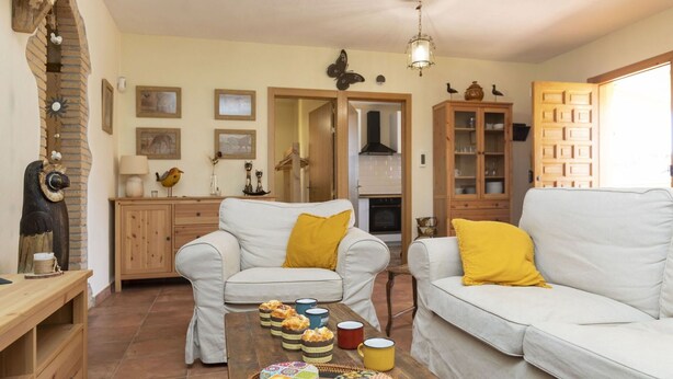 Un salon chaleureux avec deux canapés blancs, une table basse en bois sur laquelle sont disposées des pâtisseries et des tasses, un mur de briques agrémenté d’une arche décorative, et un coin cuisine visible à travers une porte ouverte.