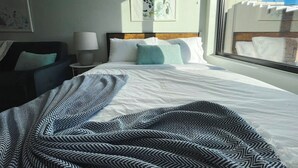 A hotel room with a large bed, a bedside table with a lamp, a window with a view of buildings, and artwork on the wall. - Sleepover | Lavish Studio and Gym - Des Moines (Des Moines)