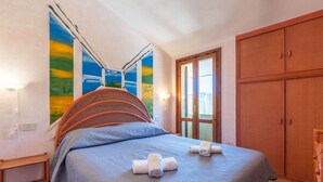 A bedroom with a bed, a wooden wardrobe, a small table, and a wall with a painting.