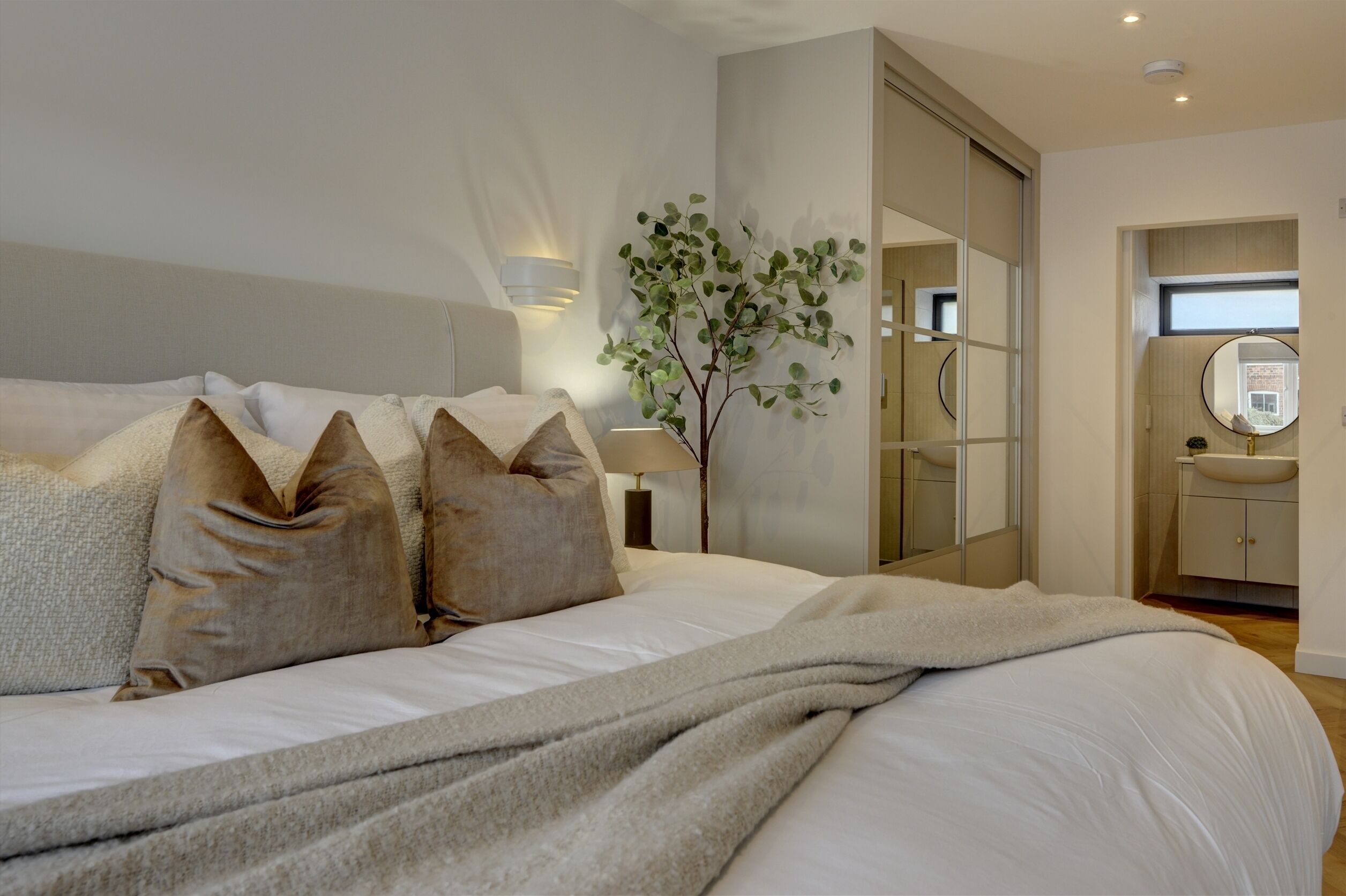 A modern bedroom with a large bed, a bedside lamp, a mirror, and a plant.