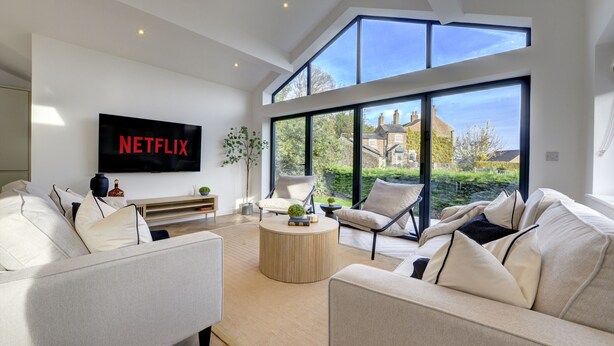 Un salon moderne doté d’un grand écran de télévision affichant le logo « NETFLIX », d’un canapé confortable, d’une table basse et d’une vue sur une maison à travers de grandes fenêtres.