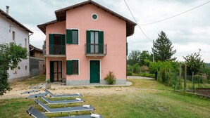 Un bâtiment à deux étages avec une façade rose, des volets verts et un balcon. Plusieurs chaises longues sont disposées dans le jardin avant.