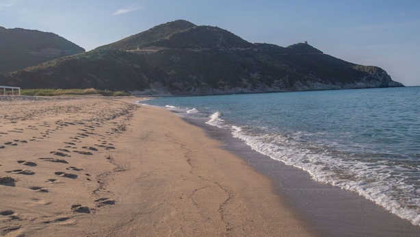 Ein Strand mit Fußspuren, die zum Wasser führen, eine weiße Struktur im Sand und ein Berg im Hintergrund.