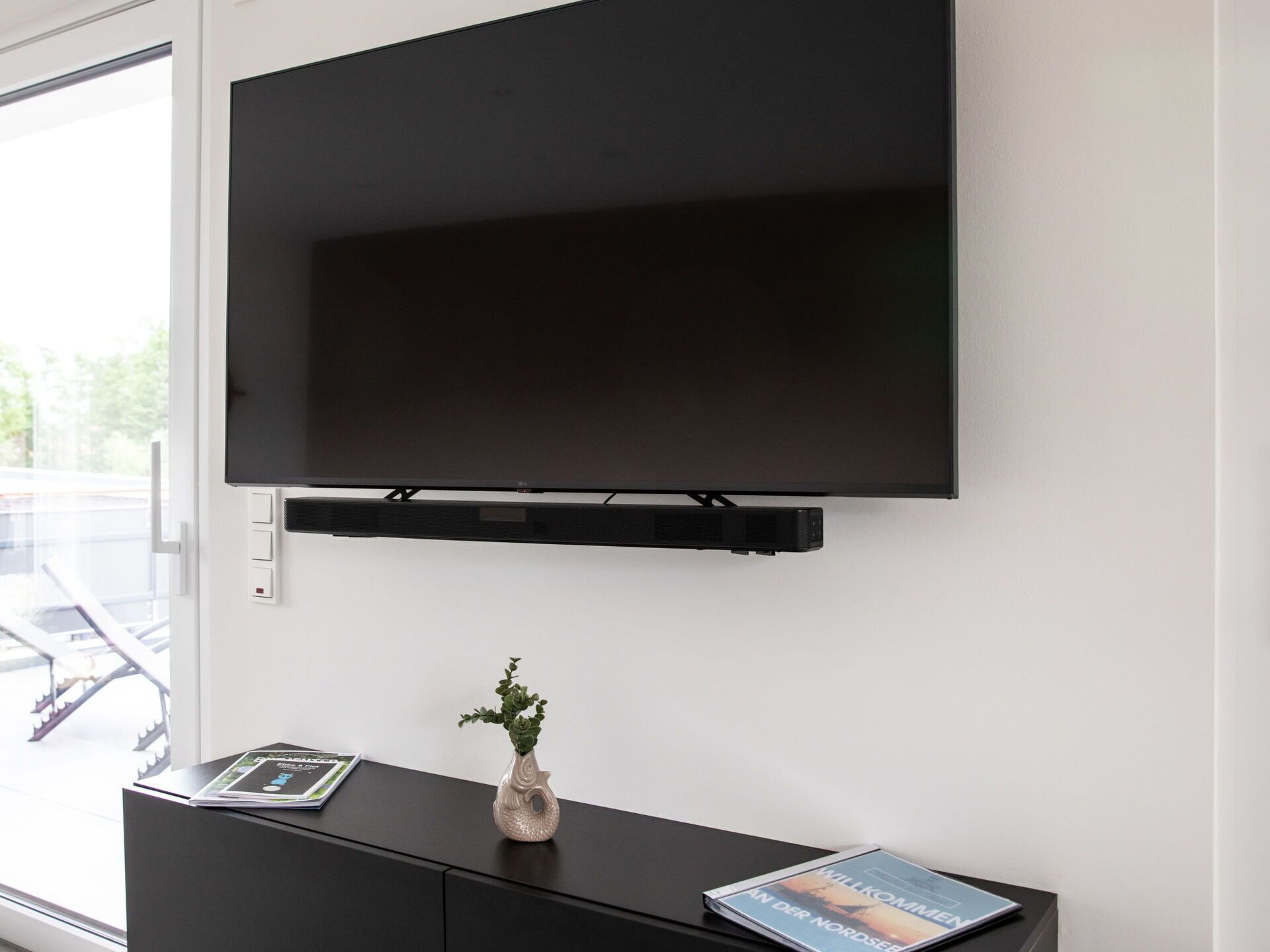Un salon moderne avec un téléviseur à écran plat fixé au mur, un meuble noir pour appareils électroniques en dessous, et une petite plante en pot posée sur le meuble.