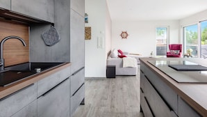 Cozinha moderna com armários cinza, bancada de madeira e fogão embutido. Há um sofá cinza com almofada vermelha na sala de estar, e uma cadeira vermelha perto de uma varanda com vista para o exterior.
