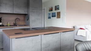 Een moderne keuken met grijze kastjes, een houten aanrechtblad en een inbouwgootsteen. In de aangrenzende zithoek staat een witte bank en een rode kussen.