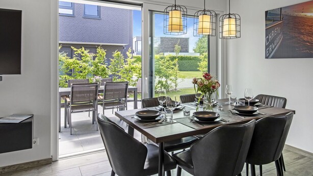 Een moderne eetruimte met een grote tafel voor zes personen, een glazen deur naar een terras buiten, en een schilderij aan de muur.