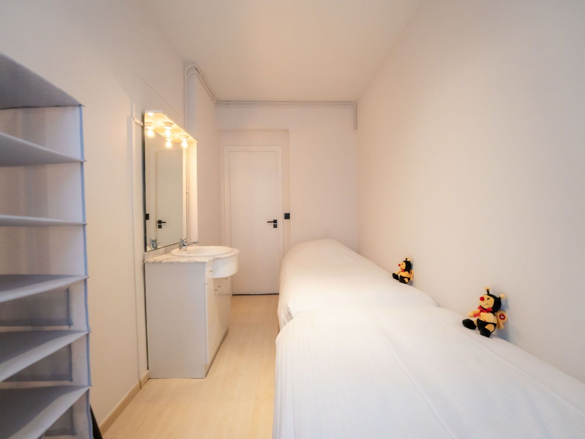 Ein kleiner, weißer, schmaler Raum mit einem Bett, einem weißen Kleiderschrank mit Regalen, einem Spiegel und einem Waschbecken.