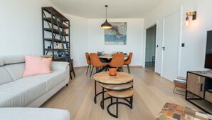 A modern living room with a sofa, a coffee table, and a dining area with chairs. There is a bookshelf and a painting on the wall.