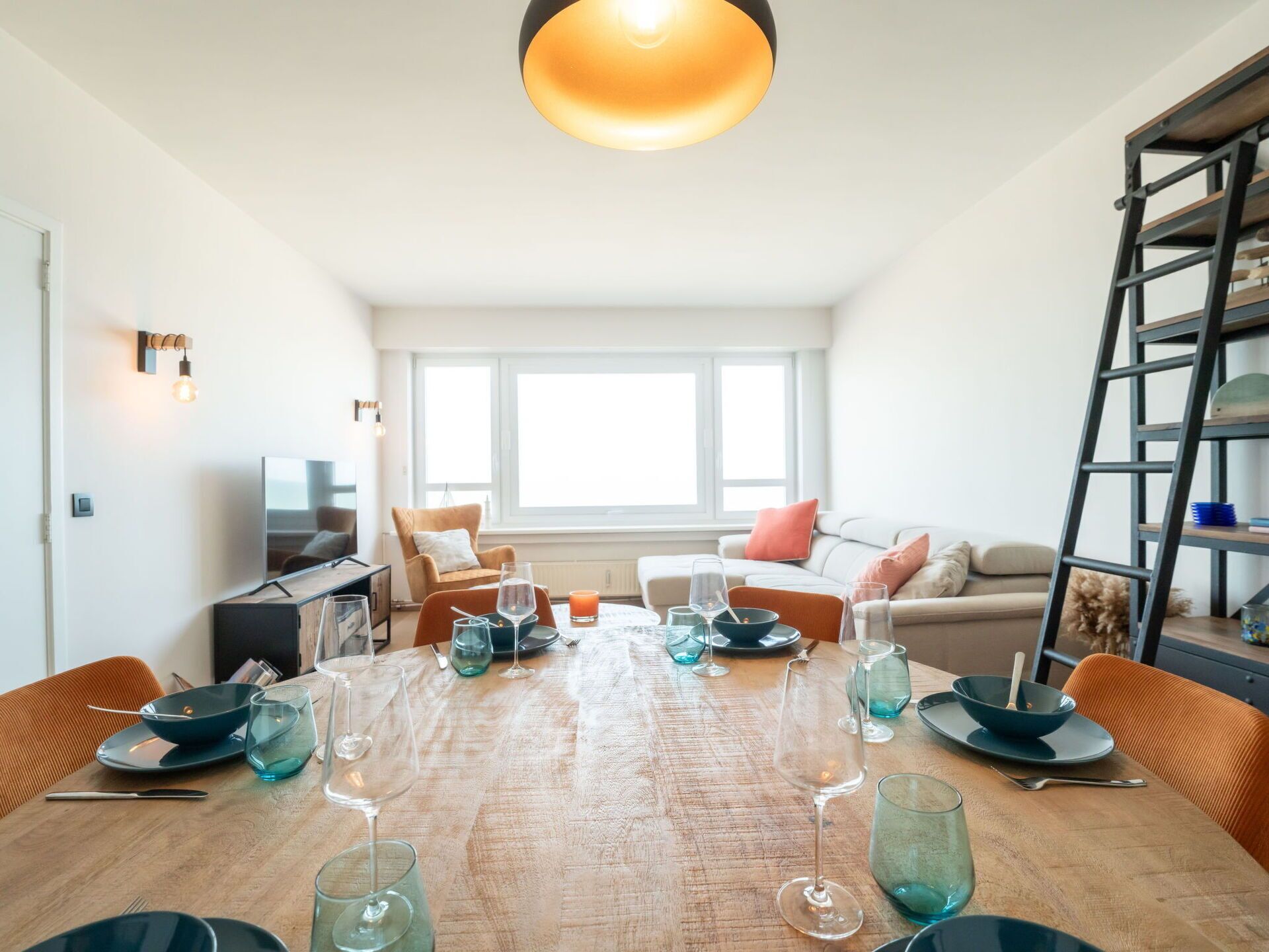 A modern living room with a wooden dining table set for a meal, a sofa, a TV, and a ladder in the corner.
