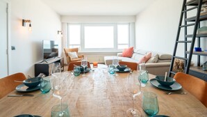 A modern living room with a wooden dining table set for a meal, a sofa, a TV, and a ladder in the corner.