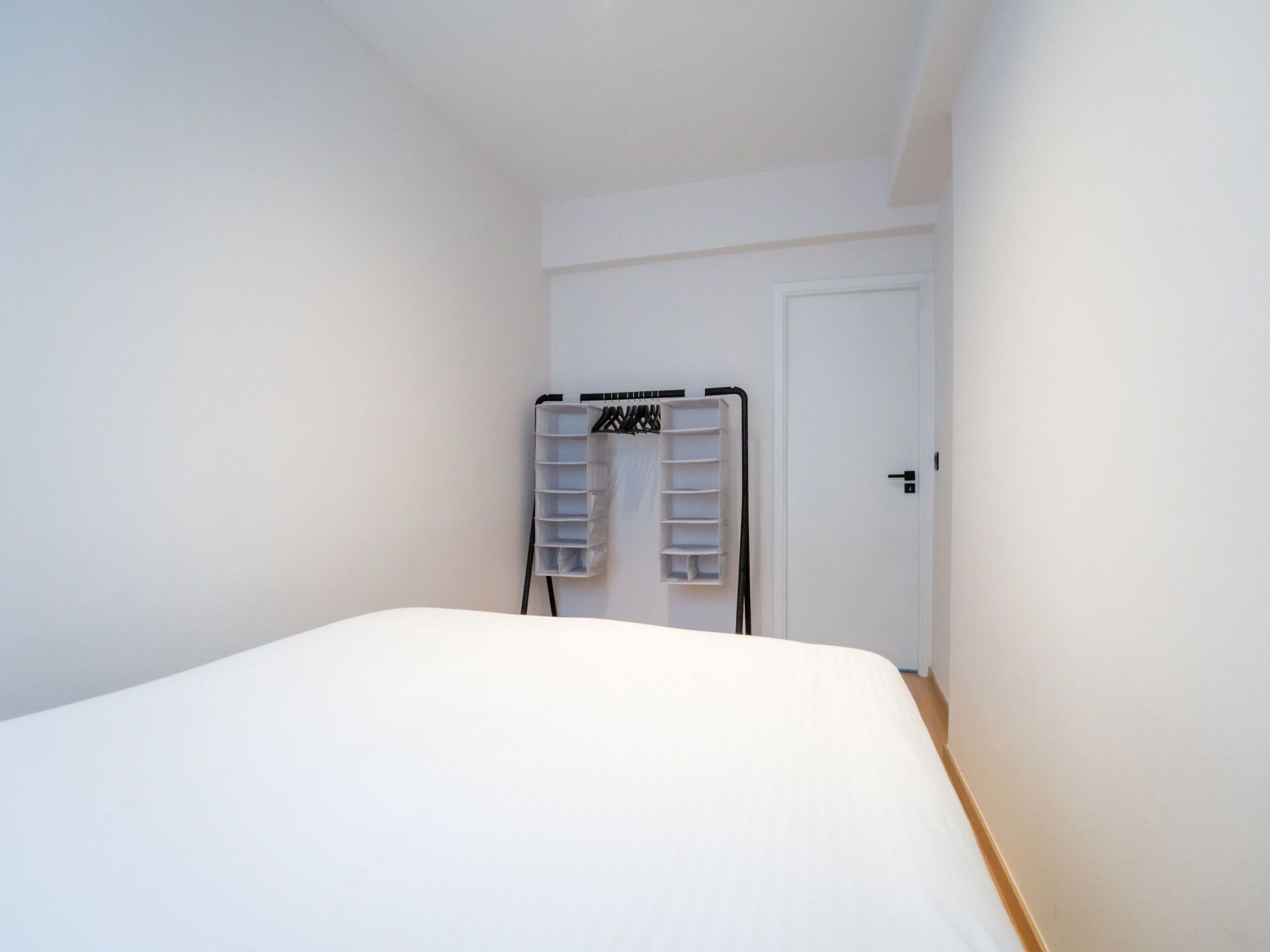 A small, clean room with a bed, a white door, and a storage unit with shelves.
