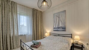 A bedroom with a bed, two bedside tables, a painting, and a window with curtains. - Cosy apartment for 3 guests with A/C, WIFI, TV, patio and panoramic view (Rijeka)