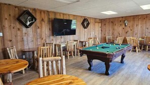 Ein Zimmer mit Billardtisch, Holzstühlen und einem an der Wand befestigten Fernseher.