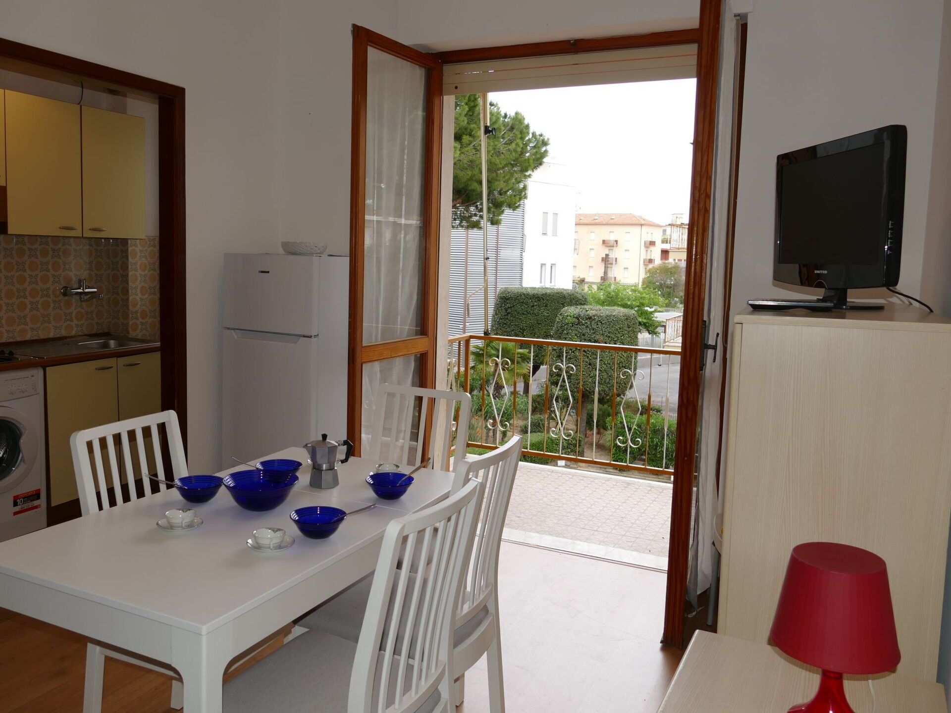 Une petite cuisine équipée d’un réfrigérateur, d’un lave-linge et d’un coin repas avec une table et des chaises. Il y a un balcon avec vue sur les immeubles et un téléviseur posé sur un meuble.