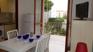Une petite cuisine équipée d’un réfrigérateur, d’un lave-linge et d’un coin repas avec une table et des chaises. Il y a un balcon avec vue sur les immeubles et un téléviseur posé sur un meuble.