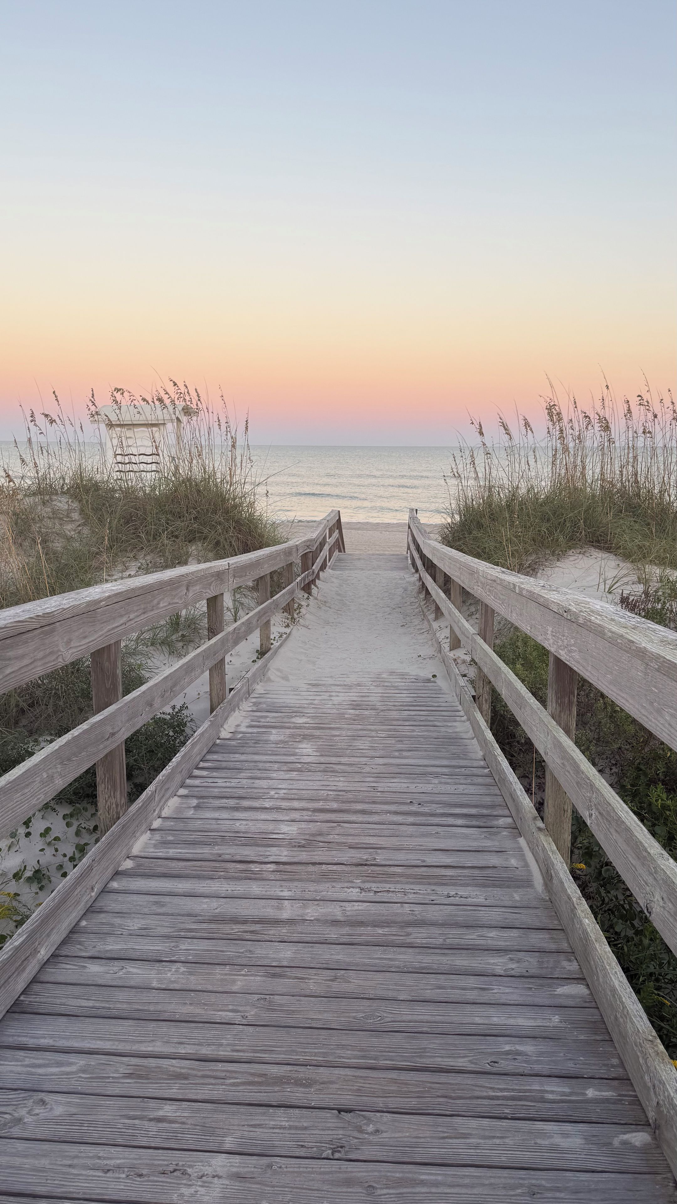A wooden boardwalk leading to a beach with sand and grass on either side.