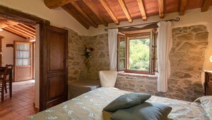Un dormitorio con pared de piedra, techo de madera, cama con ropa de cama floral, ventana con cortinas y una silla.