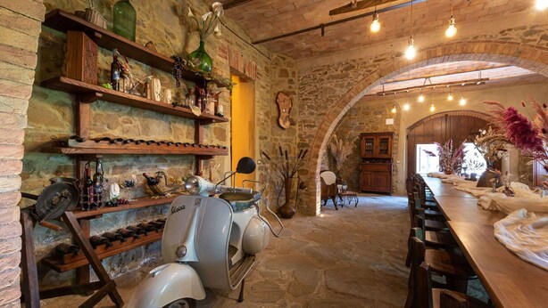 A rustic interior with stone walls, a vintage Vespa, wooden shelves with bottles, and a long wooden table. - Lovely apartment in villa with A/C, WIFI, pool, TV, patio, pets allowed and panoramic view (Cinigiano)