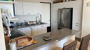 Een moderne keuken met witte kasten, een granieten aanrechtblad, een geïntegreerde koelkast en rieten stoelen.