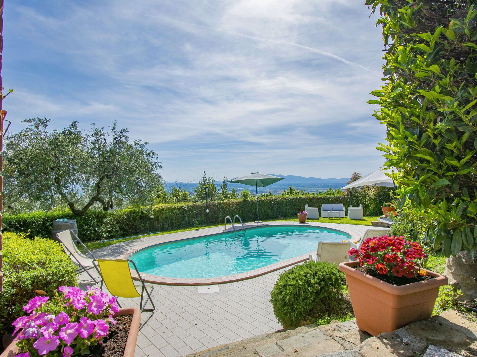 A swimming pool with a view of mountains, surrounded by greenery and outdoor furniture.