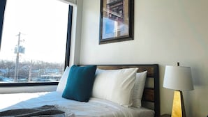 A hotel room with a bed, a nightstand, a lamp, a window with a view of buildings, and a framed picture on the wall.