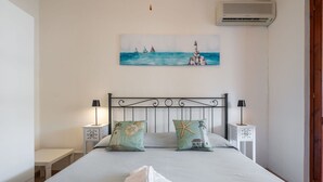 Un lit bien fait, avec deux oreillers, une table de chevet sur laquelle se trouve une lampe, et un tableau représentant des voiliers et un phare accroché au mur.