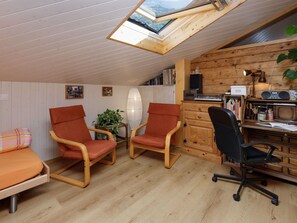 Een knus vertrek met schuine wanden, houten lambrisering, een bureau met computer, twee stoelen, een boekenkast en een dakraam.