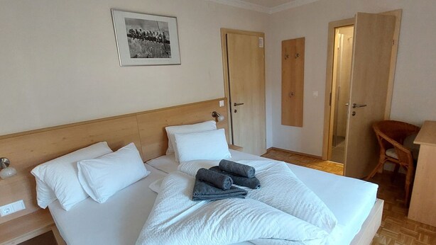 Ein ordentlich bezogenes Bett mit weißen Leinen und zusammengerollten Handtüchern, ein Holz-Kopfteil, ein gerahmtes Foto an der Wand und Holztüren.