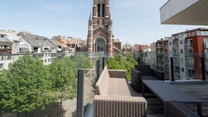 Une vue d’un clocher d’église avec une horloge, entouré de bâtiments modernes et d’un balcon agrémenté de meubles en osier.