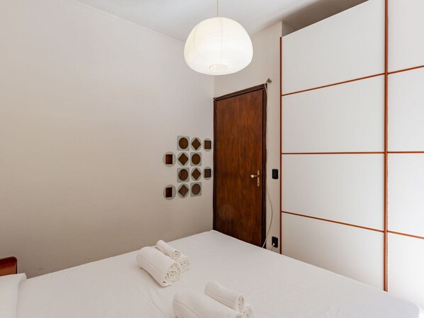 A neatly made bed with two rolled towels, a wooden wardrobe, and a wall with geometric patterns.