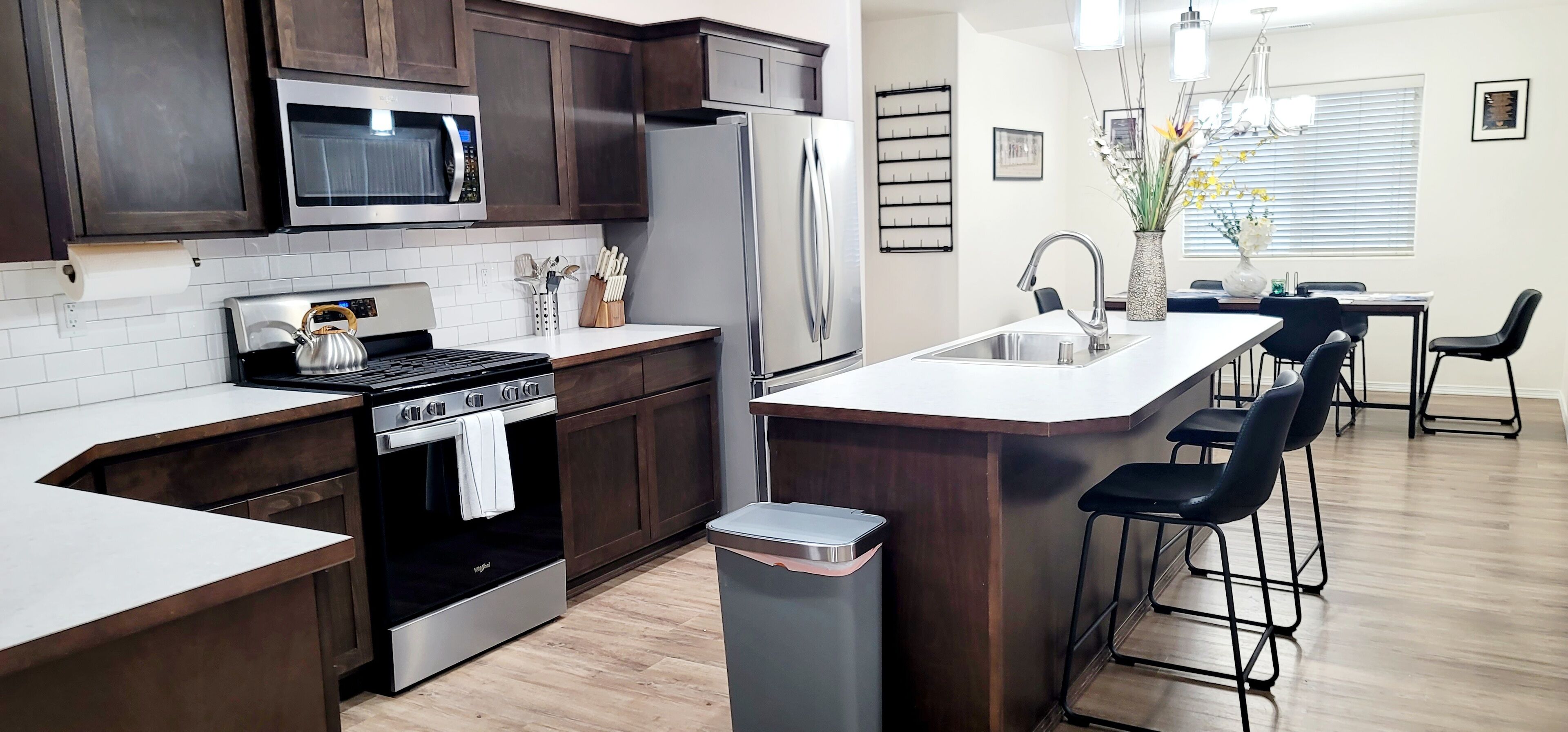 A modern kitchen with dark wood cabinets, stainless steel appliances, a white countertop, and a bar area with black bar stools.