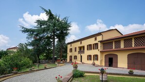 Ein Innenhof mit Kiesweg, einem großen Baum und einem gelben Gebäude mit braunen Fensterläden.