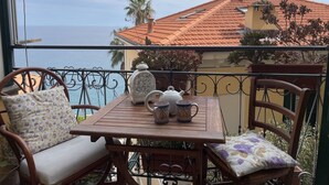 Un balcón con una mesa de madera, dos sillas y vista al mar y un edificio con tejado rojo.