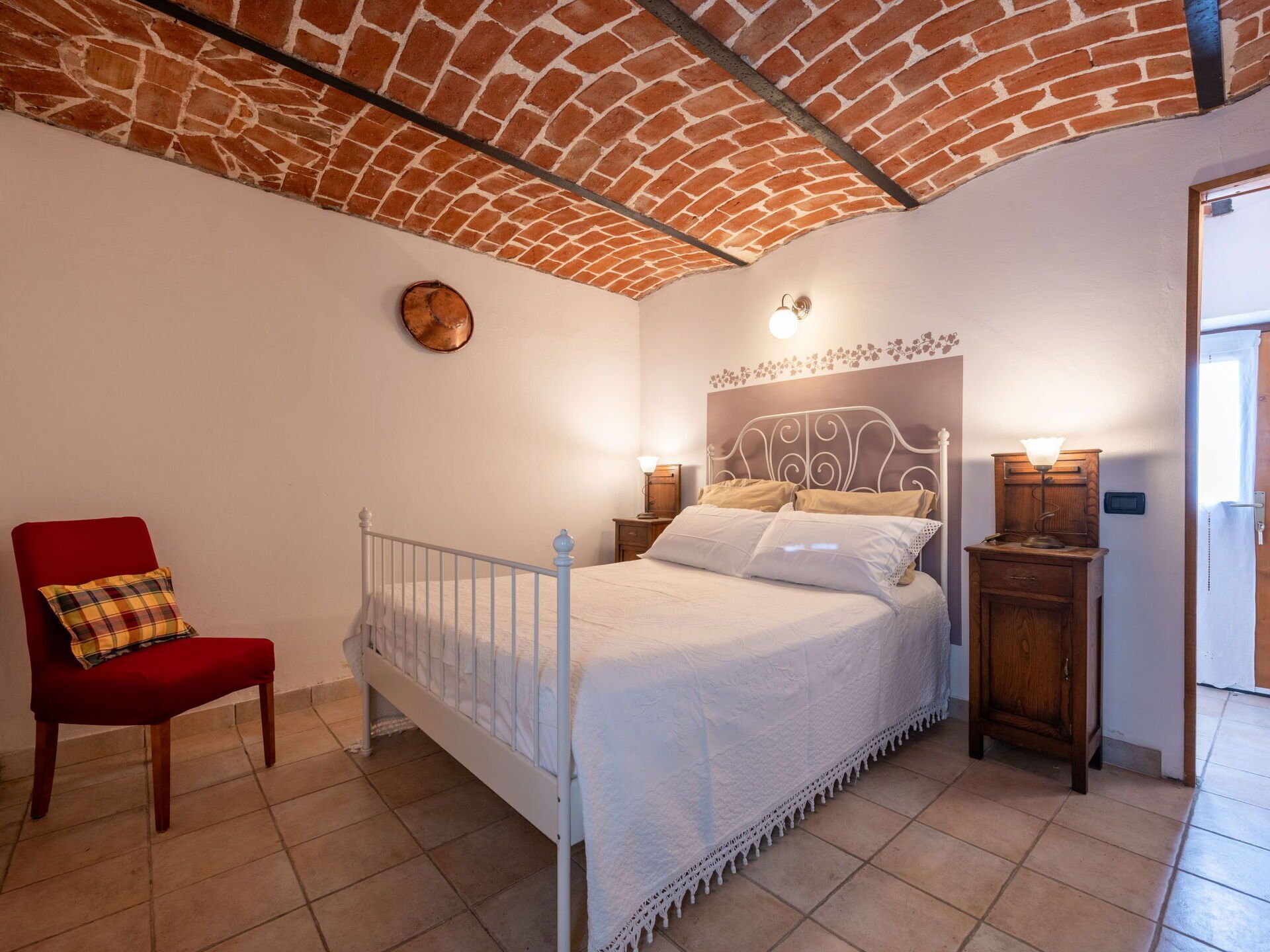 Quarto com teto de tijolos, cama branca com cabeceira decorativa, cadeira vermelha e duas mesas de cabeceira com luminárias.